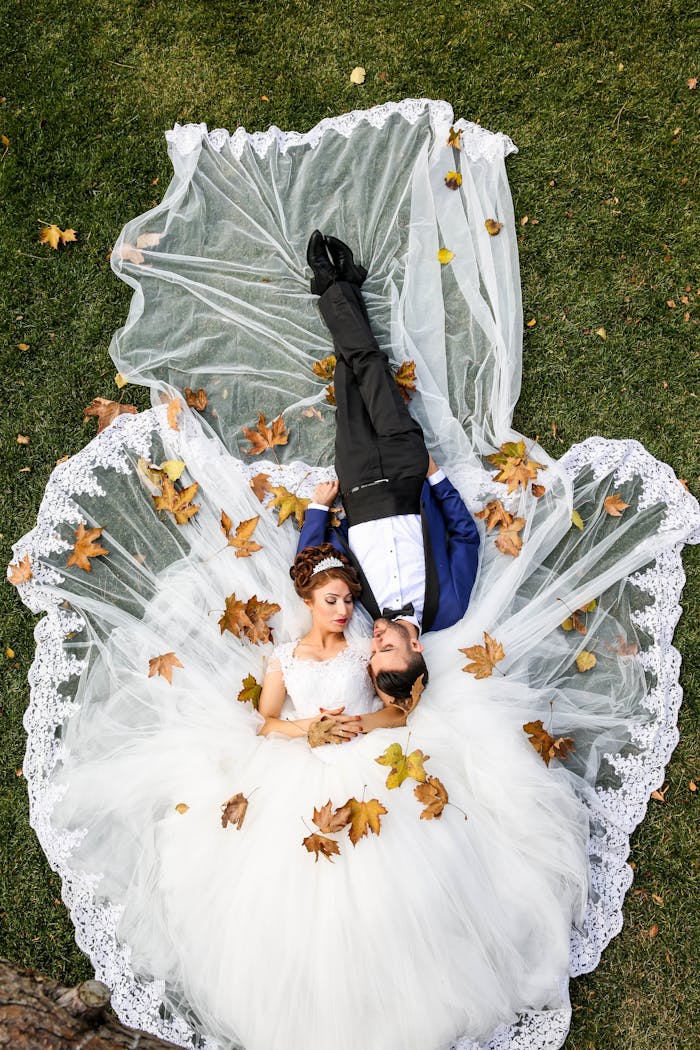 Bride and groom lying together on a lawn surrounded by autumn leaves, captured from above.